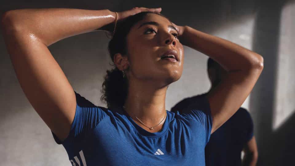 Close-up of one person in a blue workout top with arms raised behind the head, sweating after exercise. Another person in dark workout gear is visible in the background.