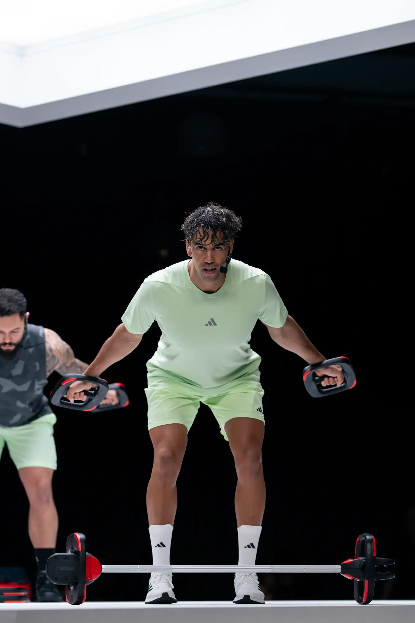 Two people exercise against a black background. One in front wears a mint green t-shirt and shorts, white socks and sneakers with black stripes, lifting weights. Another in camo top is partially visible.