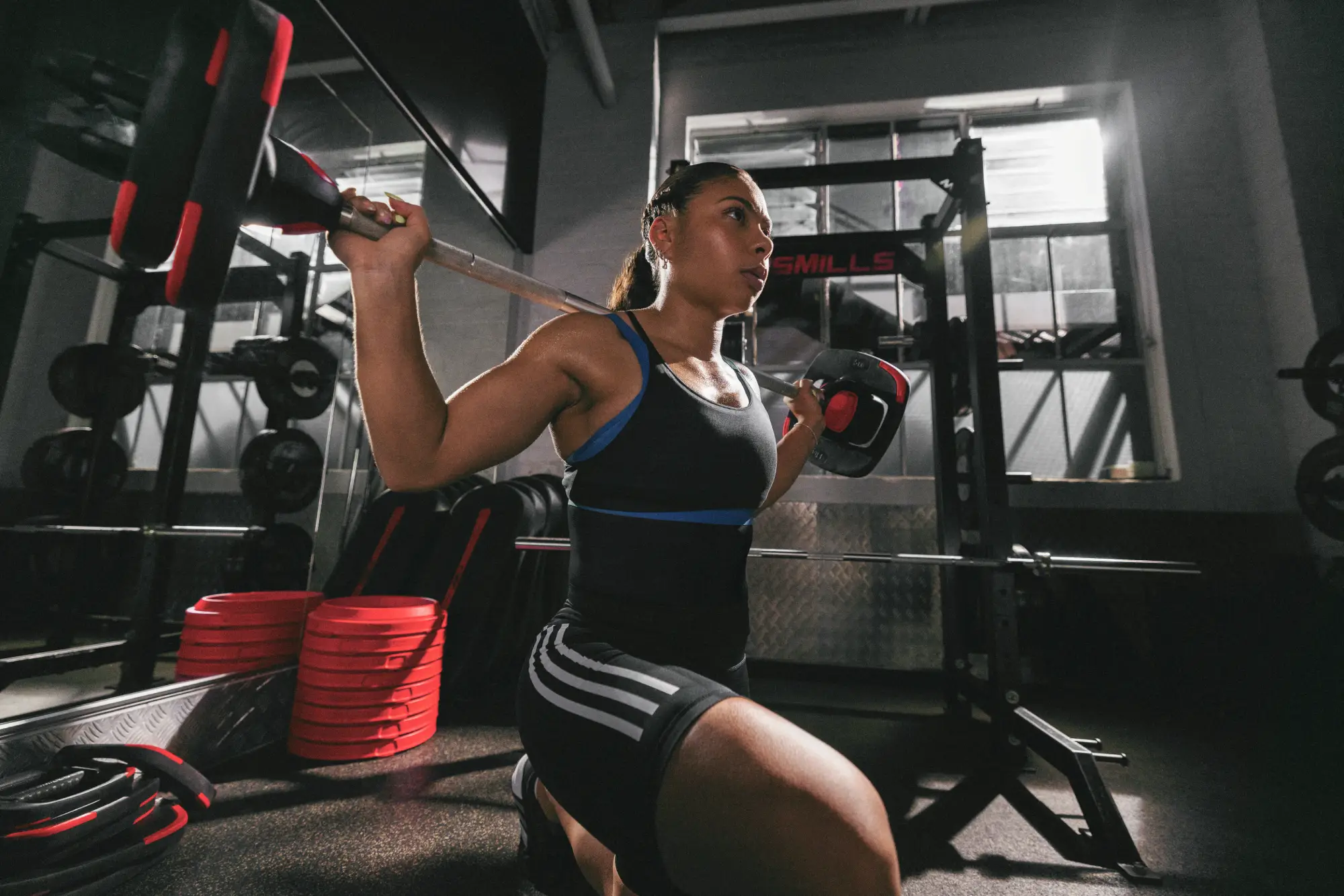 One person in a black tank top and black shorts with white stripes performs a squat with a barbell in a gym. Red and black weight plates and equipment are visible in the background.
