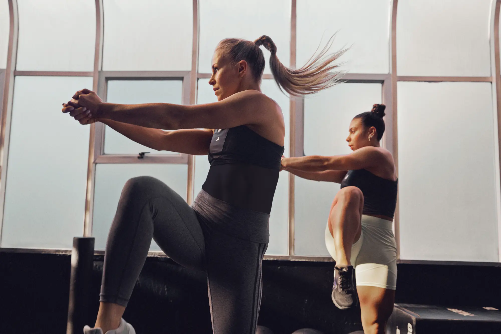 Two people exercise indoors - one in grey leggings and black top, the other in white shorts and black top - raising one knee with arms extended. Sunlight streams through large windows.