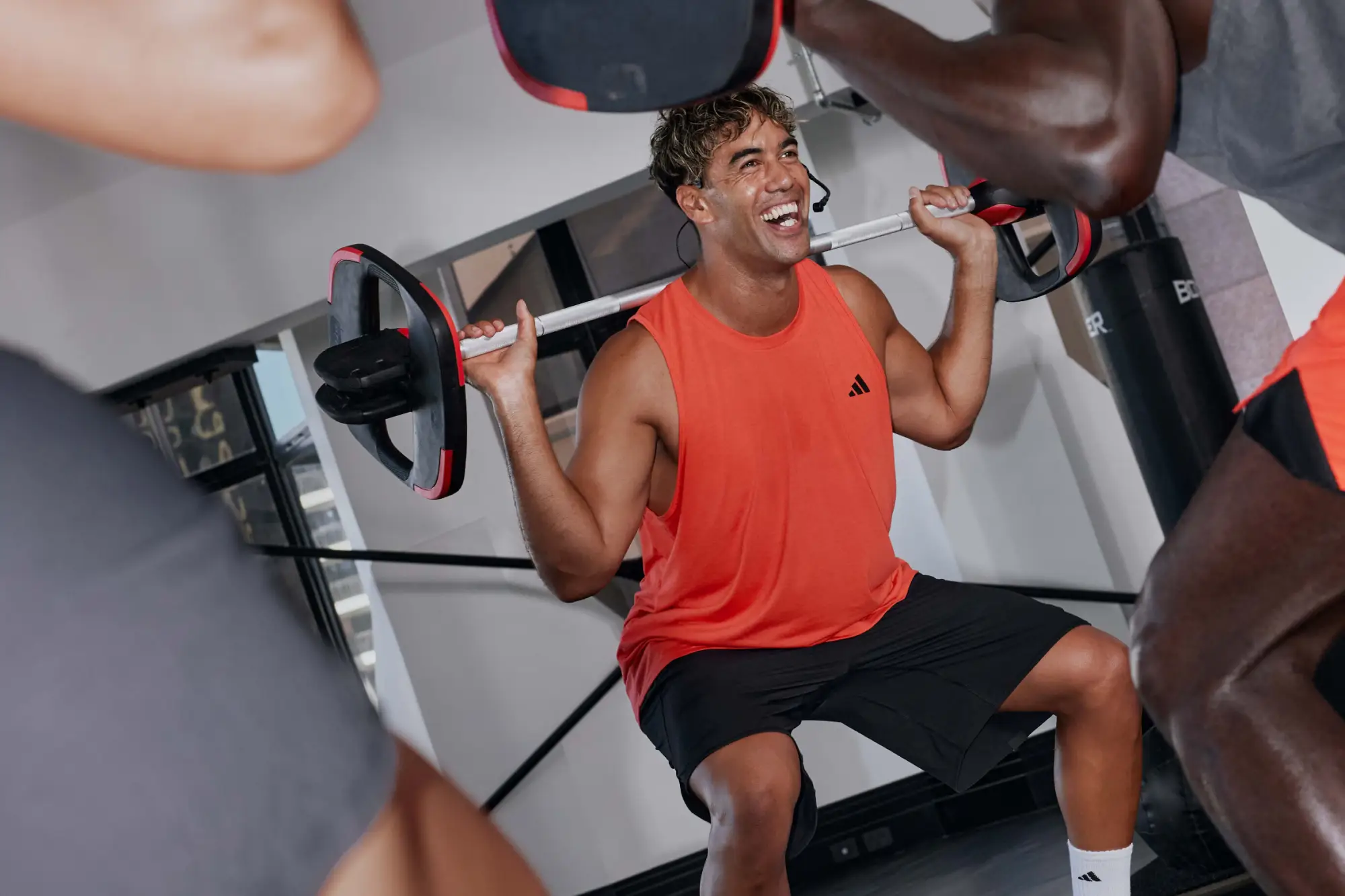 A man in an orange tank top and black shorts squats with a barbell in a gym. Three others in workout clothes are partially visible. The gym has white walls and equipment.