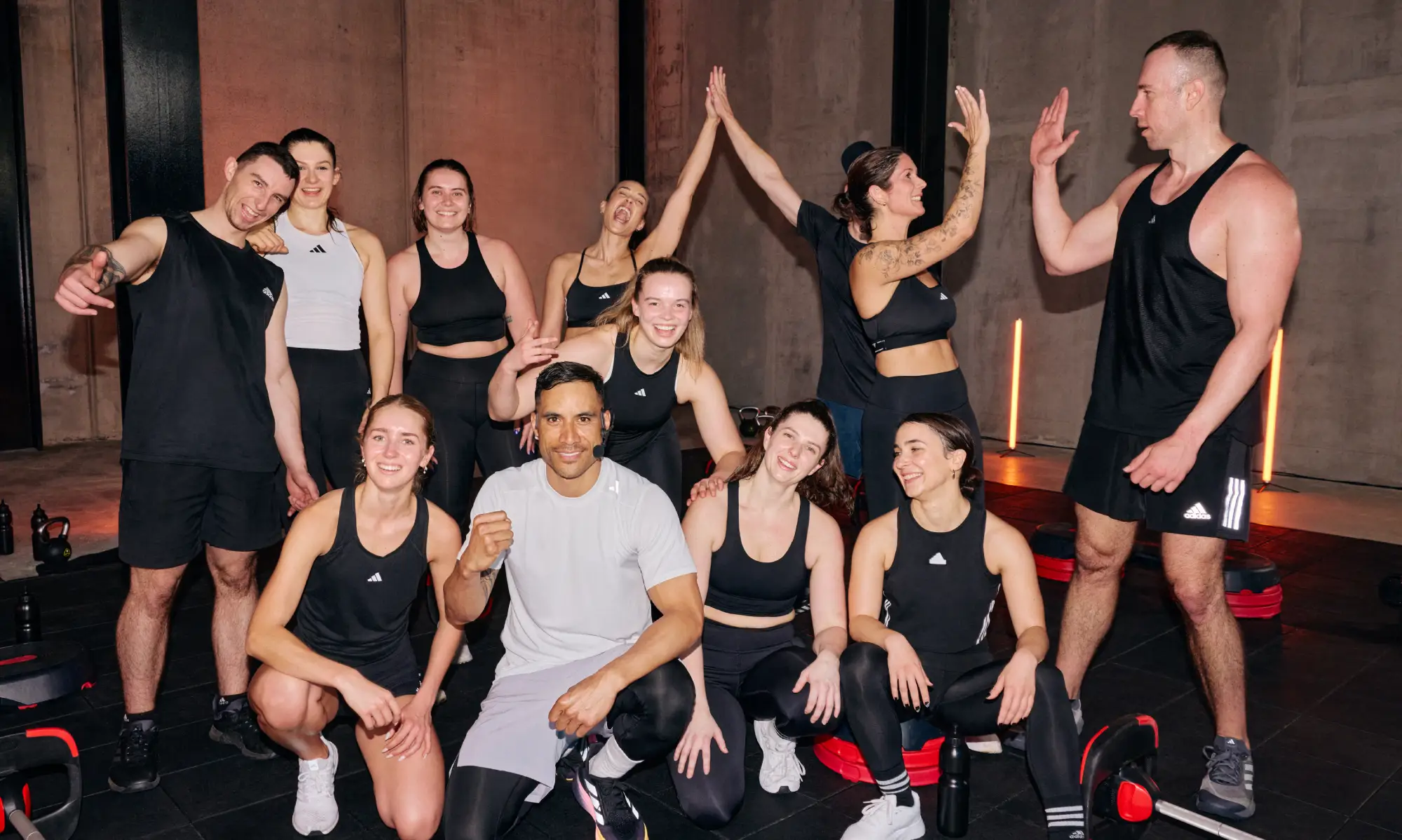 Eleven people in mostly black athletic wear with white accents pose in a gym with concrete walls and dim lighting. Some are standing, others kneeling or sitting.