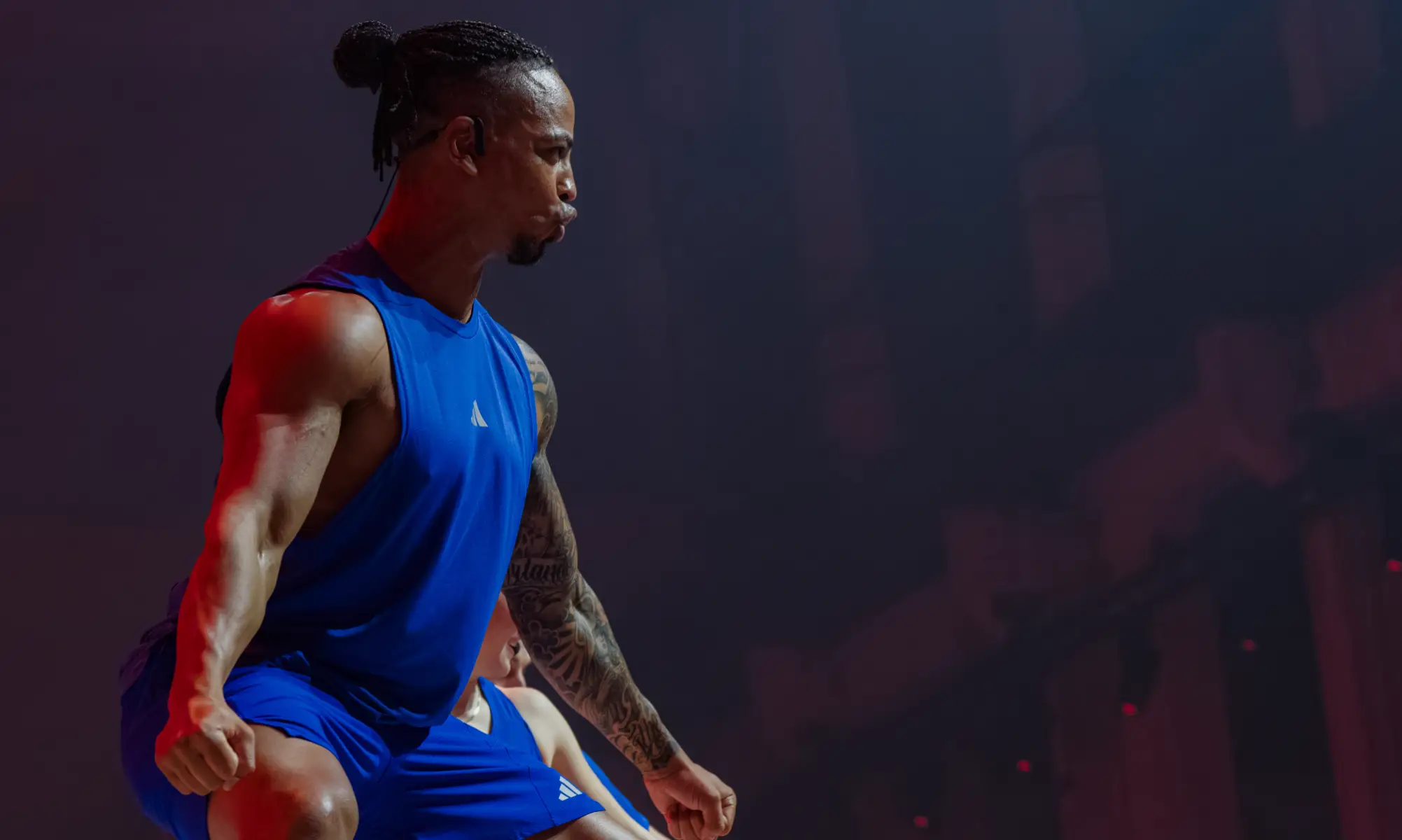 One person in a blue sleeveless shirt and shorts exercises under red lighting. Another person in similar attire is partially visible behind them.