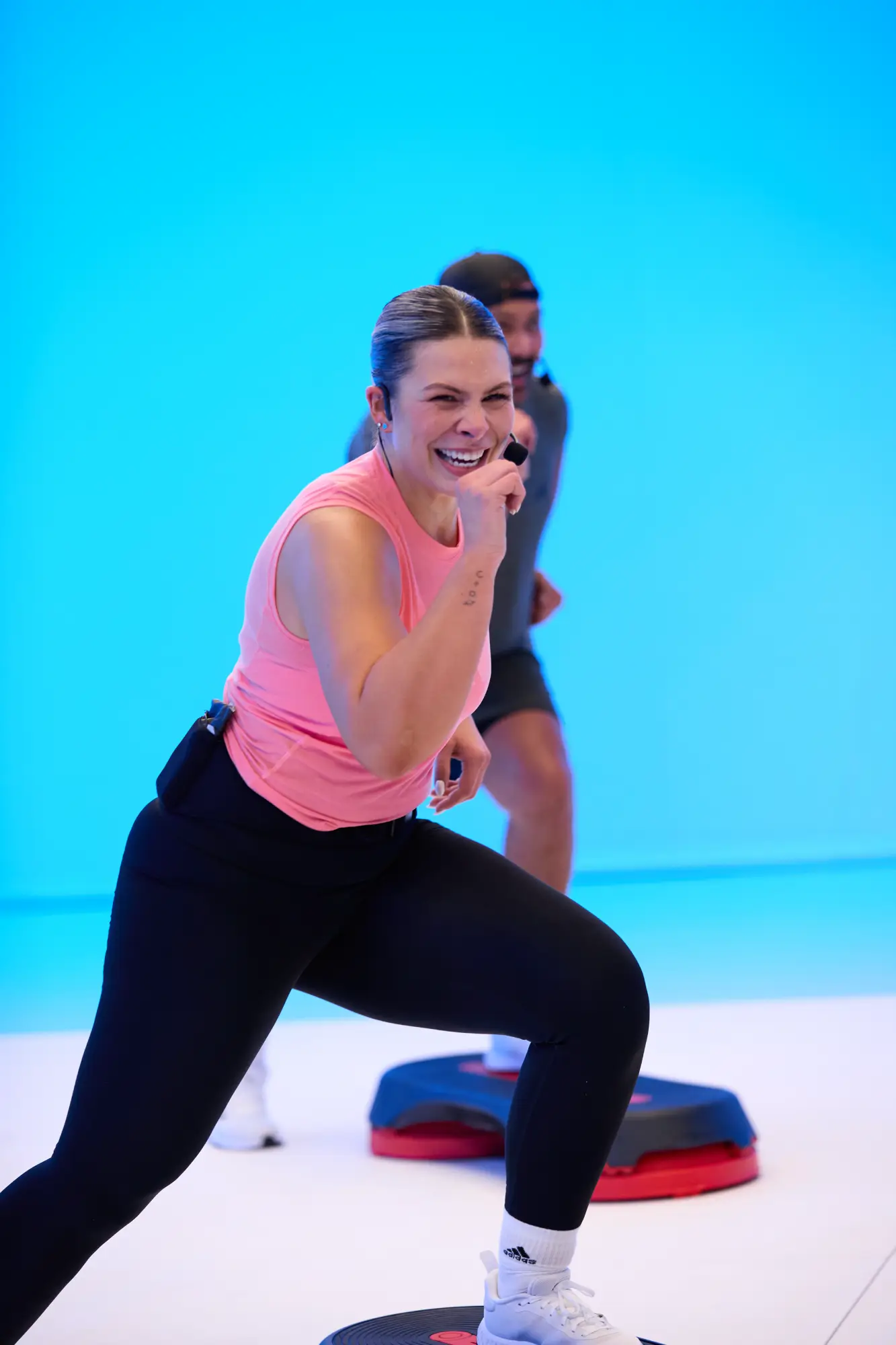 Two people in a bright room with a blue background; one in a pink top and black leggings steps on a platform, the other is partially visible in the background.