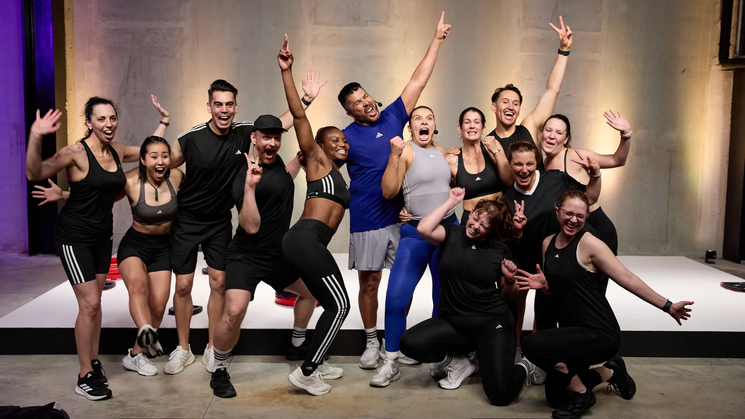 Thirteen people pose energetically in a gym; most wear black athletic wear, with one in blue and one in grey. Background features a plain wall with purple lighting on the left.