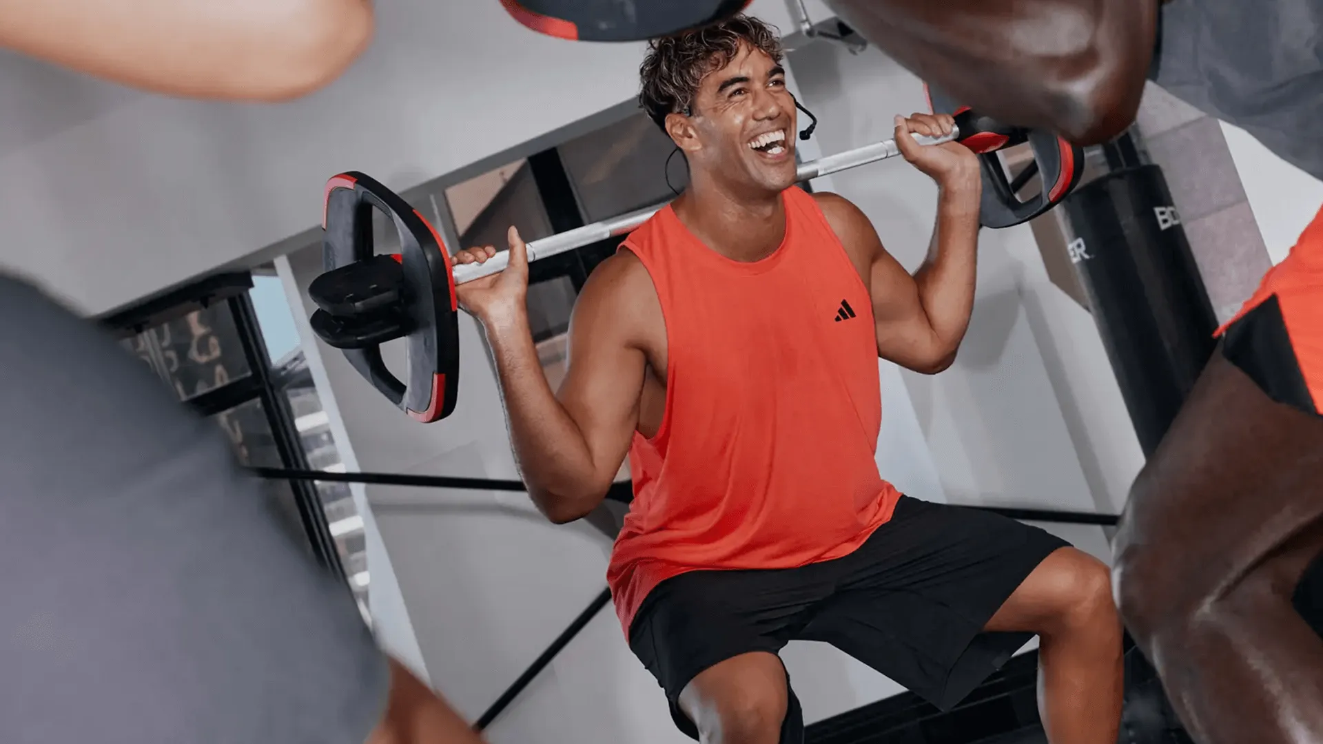 A man in an orange tank top and black shorts squats with a barbell in a gym doing Les Mills Bodypump. Three others in workout clothes are partially visible. The gym has white walls and equipment.
