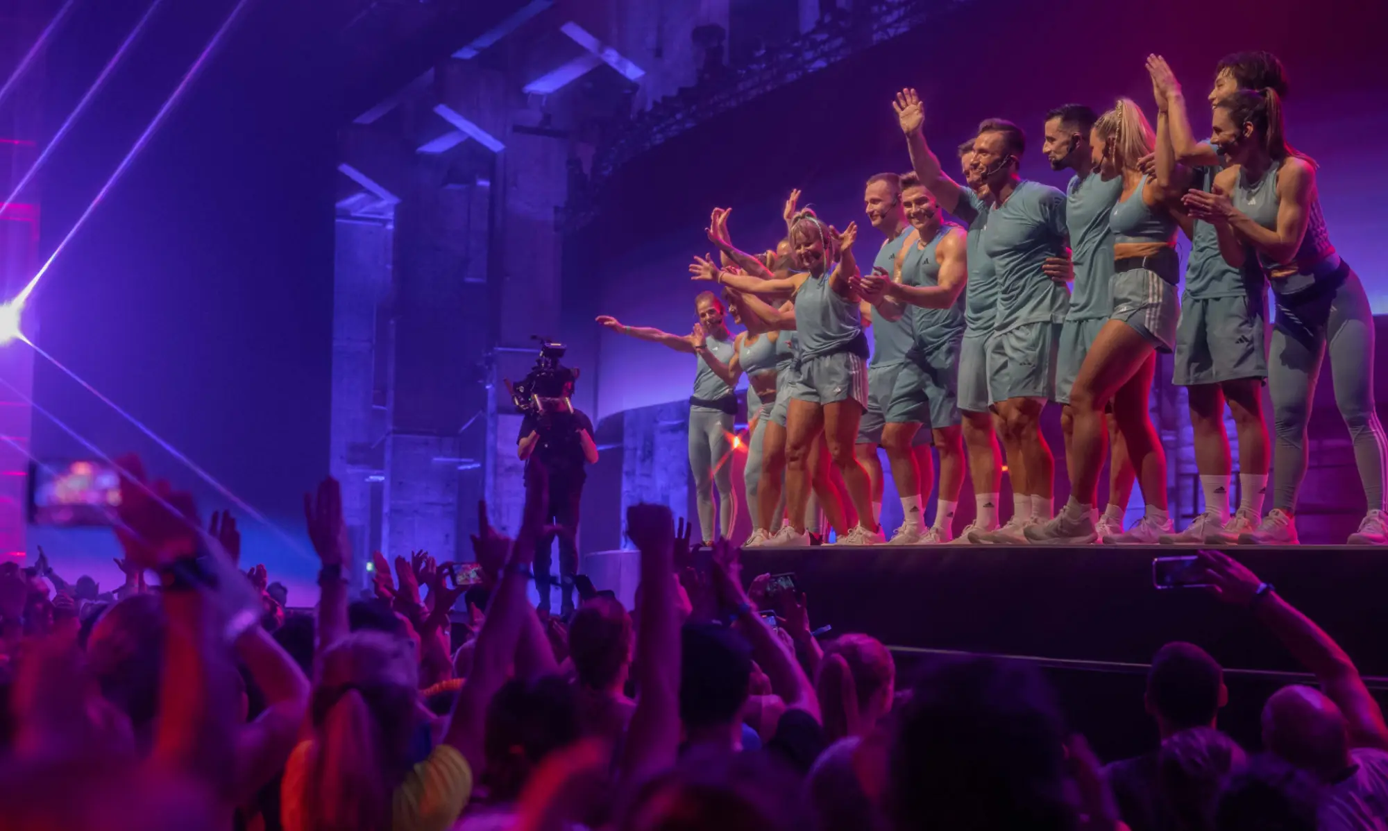 Fifteen people in light blue outfits perform on stage under purple and pink lights. A cameraman films from the left, and a cheering crowd is visible in the foreground.