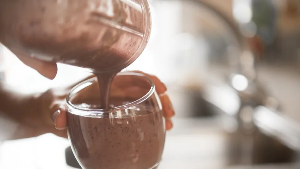 Close-up of a smoothie being poured from a blender into a round glass. The drink is purple-brown with a thick texture.
