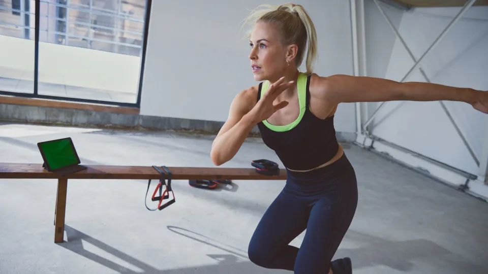 One person exercising indoors wearing a black sports bra with neon green trim and navy leggings. A wooden bench with a tablet and resistance bands is in the background.