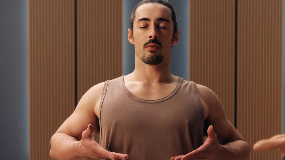 One person standing indoors wearing a sleeveless brown top, arms bent at chest level in a yoga breathing posture. Wooden panel wall in the background.