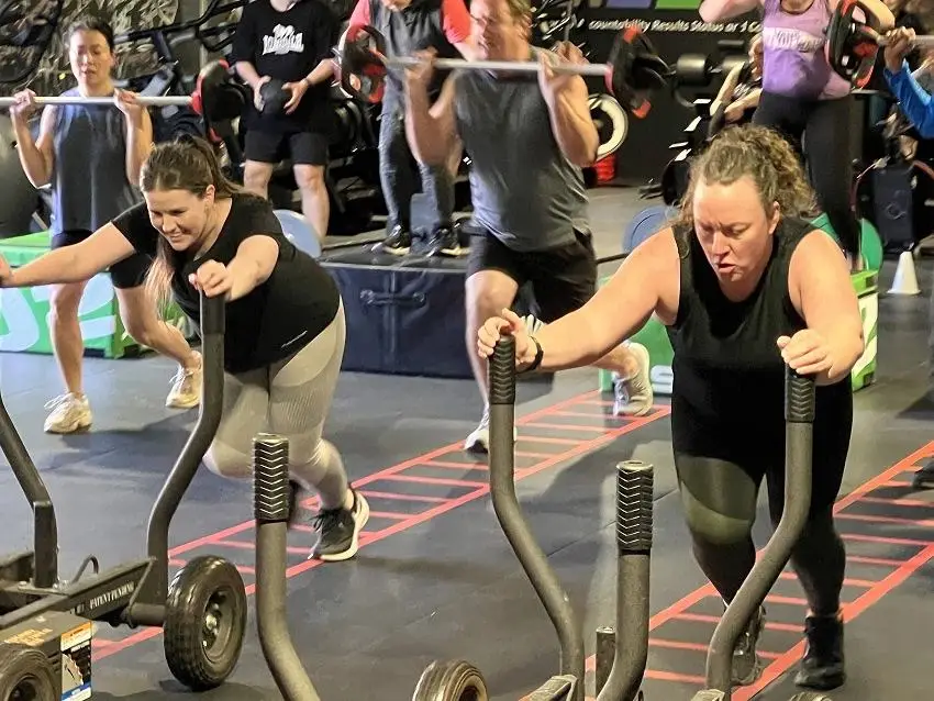 Indoor gym with 8 people exercising. Two in front push sleds on black flooring with red grid lines, others lift barbells behind. Green boxes and weight plates visible in background.