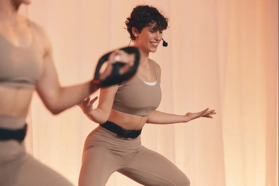 Two people in beige activewear exercising in a studio with soft peach draped background. One holds a weight plate, the other in a low stance with arms extended, wearing a black belt.