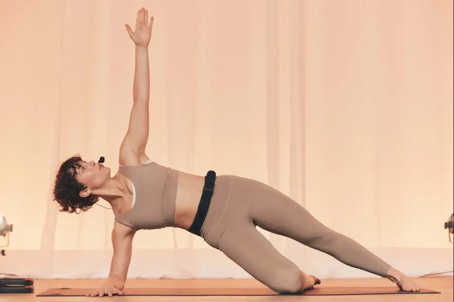 One person in beige activewear performing a side plank on a mat with one arm raised. Soft peach draped background and warm lighting create a minimal studio setting.