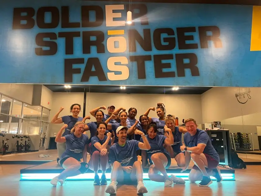 Eleven people in matching blue shirts posing in a gym with wooden flooring. Behind them is a large mirrored wall and a bold blue sign reading “BOLDER STRONGER FASTER” in black and white text.